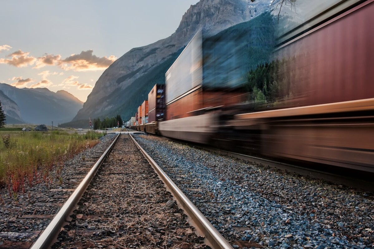A goods train with railway cars passing by quickly, creating a blur. In the distant background, there are mountains